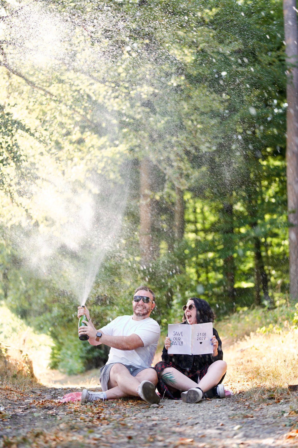 Sarah und Silvan sitzen auf einem Waldweg, Silvan lässt einen Champagnerkorken knallen, Sarah hält ein Schild „Save our Date – 21.08.2027“.